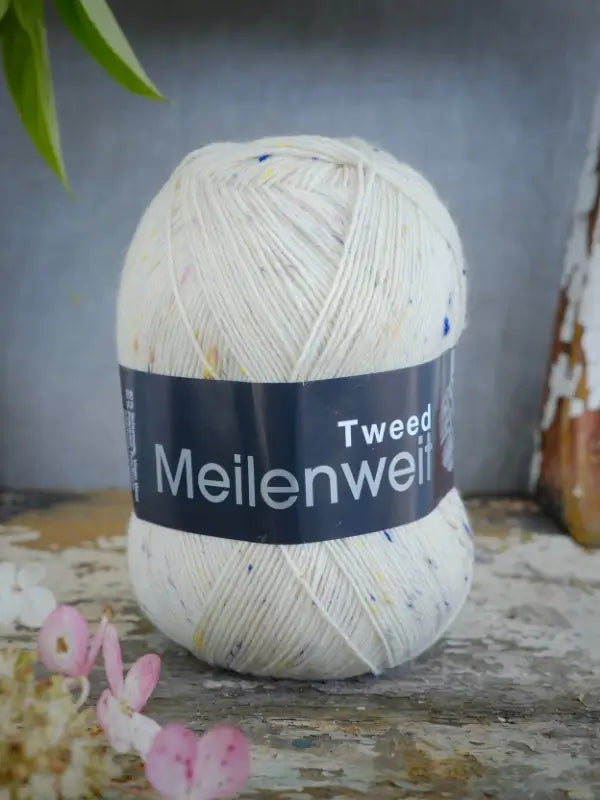 A ball of Lana Grossa Meilenweit 100 Tweed yarn, in an off-white shade with colorful tweed flecks, rests on a rustic surface beside delicate pink flowers and fresh greenery. This gorgeous sukkalanka brings charm and texture to any knitting project.