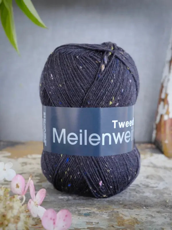 A skein of Lana Grossa Meilenweit 100 Tweed yarn in dark blue, featuring colorful flecks, rests on a rustic surface surrounded by flowers and greenery.