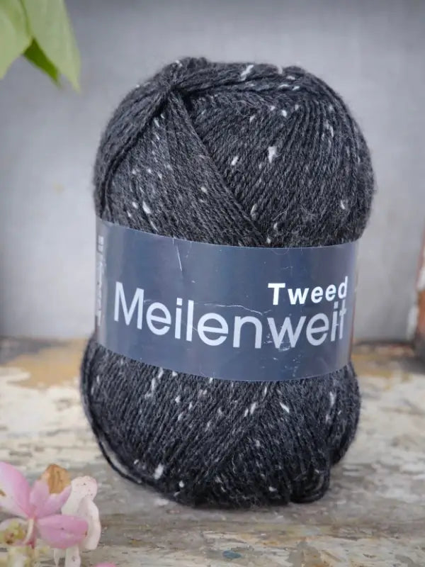 A skein of Lana Grossa Meilenweit 100 Tweed yarn in dark gray with white flecks rests on a rustic surface, accompanied by a small pink flower in the bottom left corner.