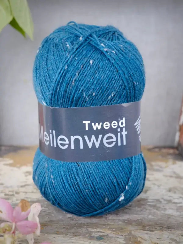 A skein of Lana Grossa Meilenweit 100 Tweed yarn in blue, adorned with delicate white flecks, rests on a rustic wooden surface. The soft yarn is encircled by its black label, while a blurred pink flower appears in the corner of the image.