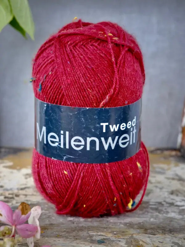 A skein of vibrant red Lana Grossa Meilenweit 100 Tweed yarn stands upright on a rustic wooden surface beside a pink flower, making it an ideal choice for your next sukkalanka project.