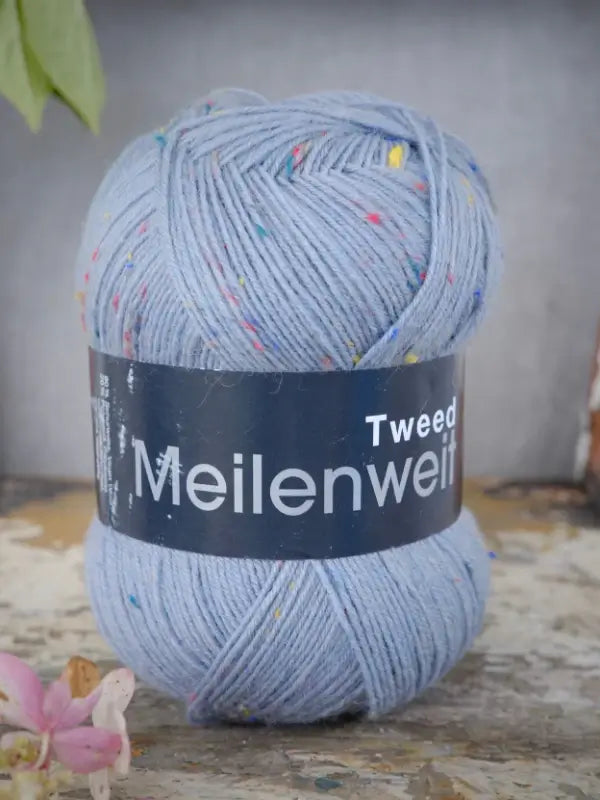 A skein of Lana Grossa Meilenweit 100 Tweed yarn in light blue with colorful specks rests on a rustic surface, accompanied by a pink flower nearby.