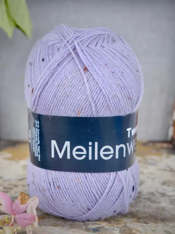 A skein of Lana Grossa Meilenweit 100 Tweed yarn in light purple with brown flecks rests on a textured surface, while a small pink flower in the bottom left corner adds a charming touch to this lovely yarn.