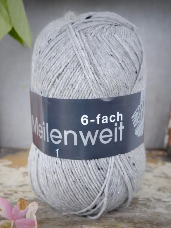 A light gray skein of Lana Grossa Meilenweit 6 Tweed yarn stands upright on a rustic surface, complemented by a flower petal in the foreground and a partial leaf at the top left. This six-ply sock yarn features subtle tweed flecks, enhancing the texture and character of the scene.