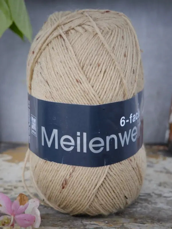 A skein of Lana Grossa Meilenweit 6 Tweed yarn in beige is placed on a rustic surface, accompanied by a pink flower and green leaves in the background, showcasing the appeal of this six-ply sock yarn.