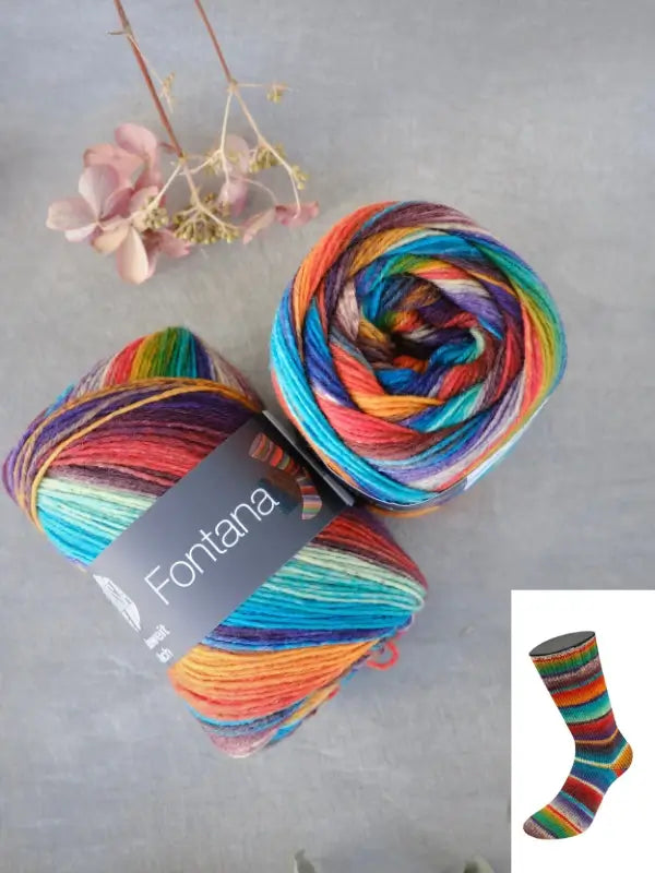 Two skeins of Lana Grossa Meilenweit 100 Fontana multicolored yarn, featuring a small image of a striped sock on the label and styled with a dried flower, are arranged on a gray surface. This high-quality sukkalanka is ideal for your next vibrant sock project.