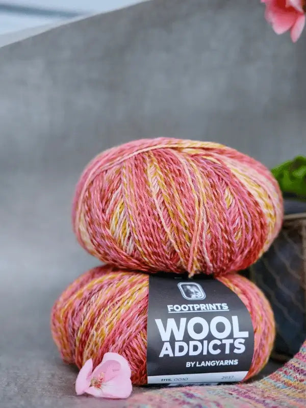 Two balls of multicolored yarn in pink, orange, and yellow shades labeled "Lang Yarns Wool Addicts Footprints" are stacked on a surface with pink flower petals nearby and part of a potted plant in the background.