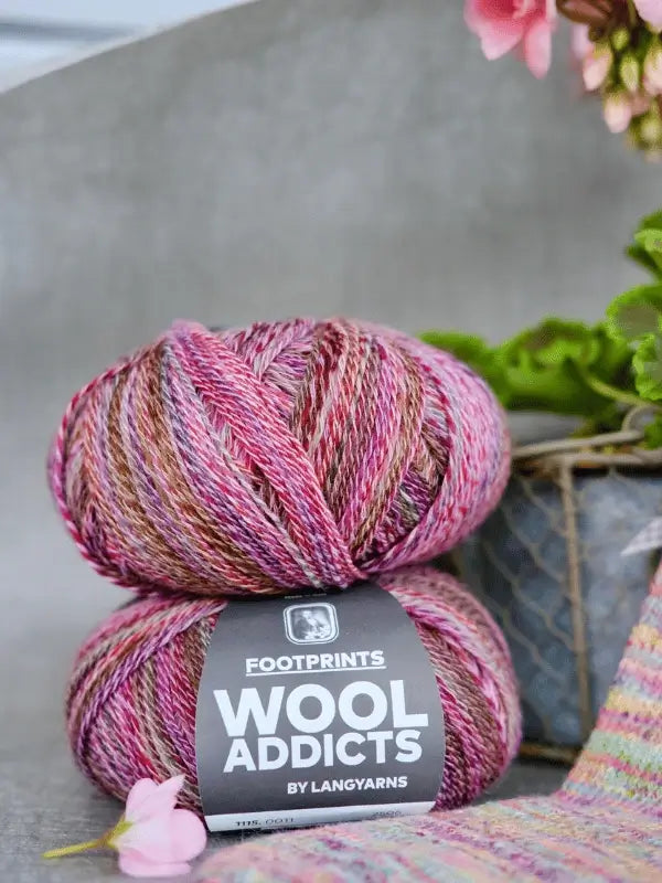 Two skeins of Lang yarns Wool Addicts footprints sukkalanka in pink, brown, and cream are stacked on a surface. A plant and a knitting project can be seen in the background.
