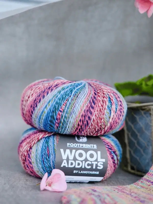 Two skeins of Lang Yarns Wool Addicts Footprints in pink, blue, and cream lie on a gray surface by a metal plant pot. The label reads "FOOTPRINTS WOOL ADDICTS BY LANGYARNS." A pink flower and soft sukkalanka add a cozy touch.