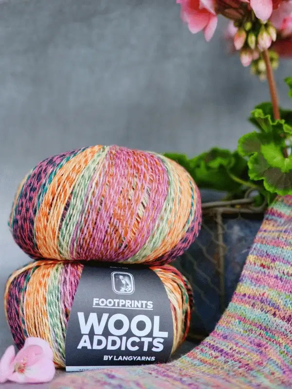 Two skeins of Lang Yarns Wool Addicts Footprints multicolored yarn are stacked beside a knitted swatch and a metal basket decorated with green leaves and pink flowers.