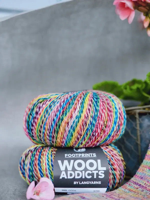 Two skeins of Lang Yarns Wool Addicts Footprints yarn are stacked together, with a pink flower and a potted plant with green leaves and pink flowers adding a charming touch beside the yarn.