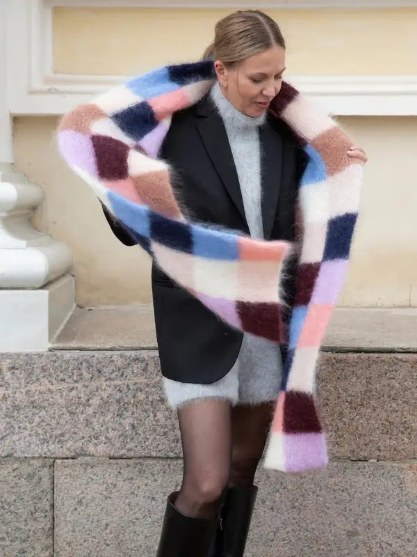 A woman in a black blazer, gray sweater dress, and black boots stands on granite steps, holding a large, colorful checkered scarf in pastel and dark shades—capturing the Nordic Chic spirit celebrated in Leeni Hoimela’s “Skandityyli: Ajattomia ohjeita nykyneulojalle.”.