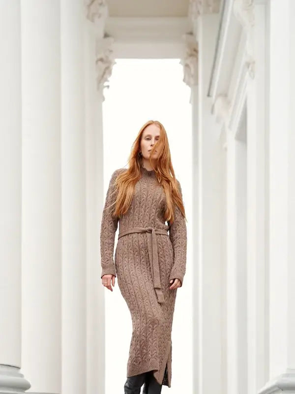 A woman with long red hair wearing a brown knit sweater dress walks between white columns, perfectly capturing the essence of Skandityyli as featured in Leeni Hoimela’s “Skandityyli, Ajattomia ohjeita nykyneulojalle,” her confident gaze reflecting timeless Nordic chic.