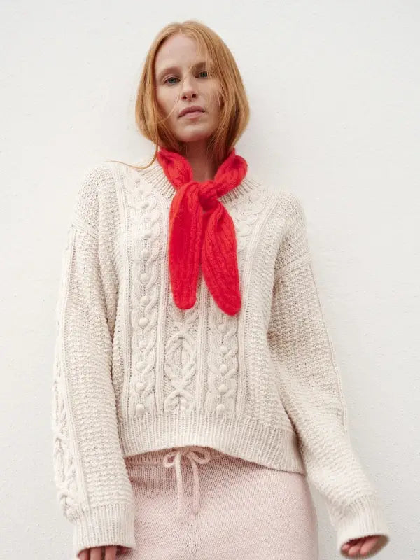 A person with long red hair wears a cozy cream cable knit sweater, matching knit pants, and a vibrant red scarf tied loosely around their neck, perfectly capturing the Nordic Chic look as featured in Leeni Hoimela’s *Skandityyli, Ajattomia ohjeita nykyneulojalle*, while standing against a simple light-colored background.