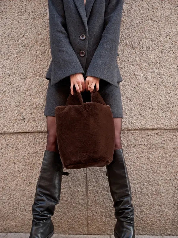 A person stands against a textured stone wall, embodying effortless Nordic Chic while holding “Leeni Hoimela – Skandityyli, Ajattomia ohjeita nykyneulojalle,” dressed in a gray blazer and matching skirt with tall black boots and carrying a large, fuzzy brown handbag.