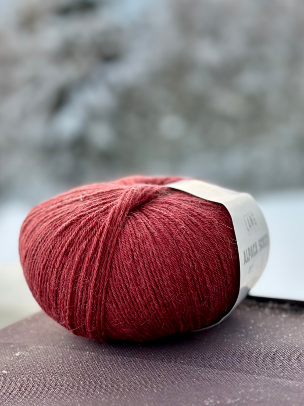 A close-up of Lang Alpaca Soxx 4ply yarn, featured in the Mittums lapasten ohje (PDF) kit, with a white label resting on a dark surface. The background is blurred and looks like a muted outdoor setting.