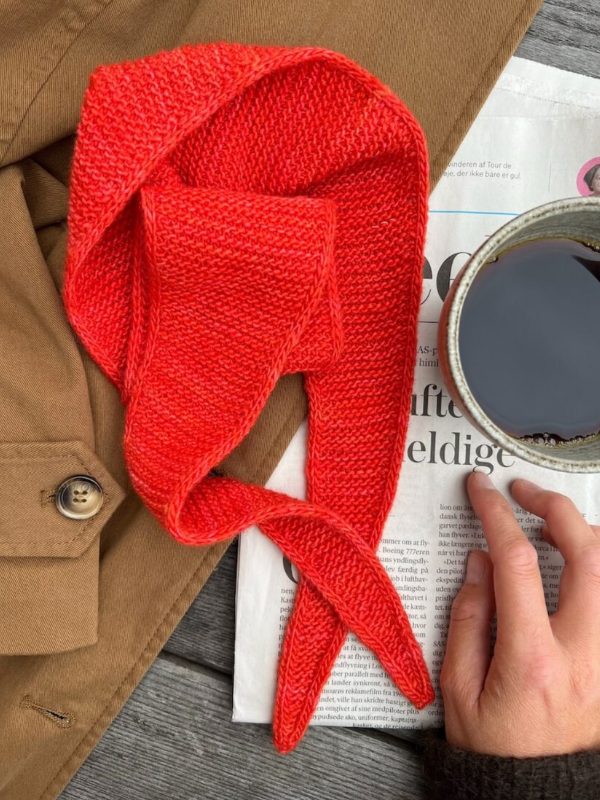 A hand holds a cup of coffee over a newspaper next to a brown coat topped with the PetiteKnit - Sophie Scarf, a bright red neulottu huivi neatly folded.