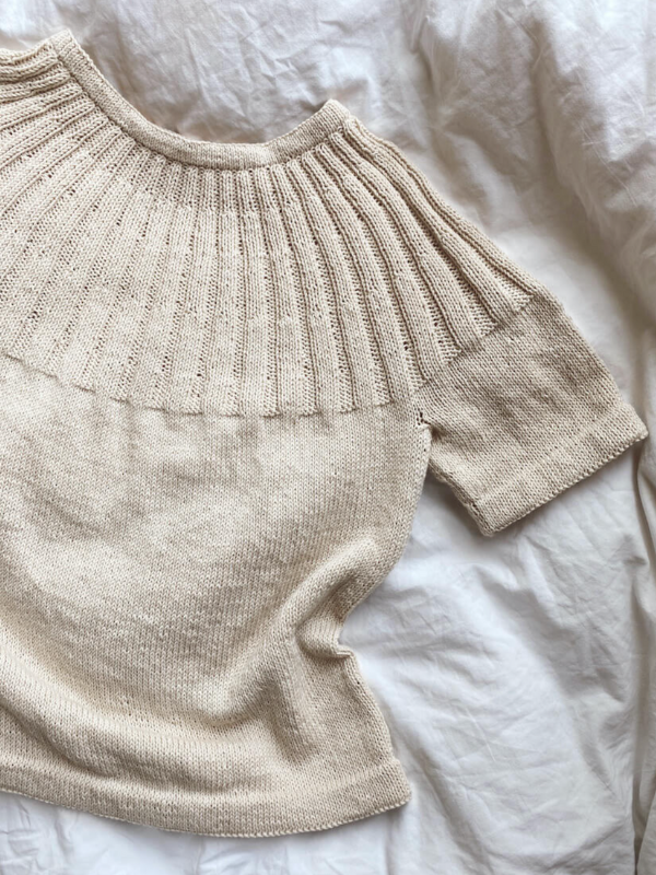 The PetiteKnit - Sunday tee neuleohje is a beige short-sleeve knitted sweater with a ribbed wide yoke, crafted top down, shown laid flat on crumpled white fabric.