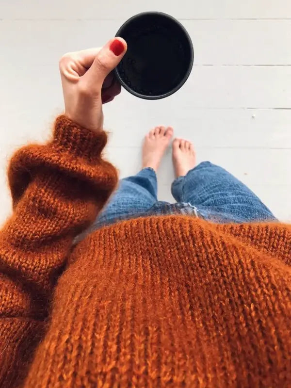 A person in a fuzzy orange PetiteKnit - Novice Sweater neuleohje and blue jeans holds a black coffee cup, their bare feet visible on a light-colored floor, viewed from above—a perfect inspiration for anyone seeking beginner-friendly knitting patterns.