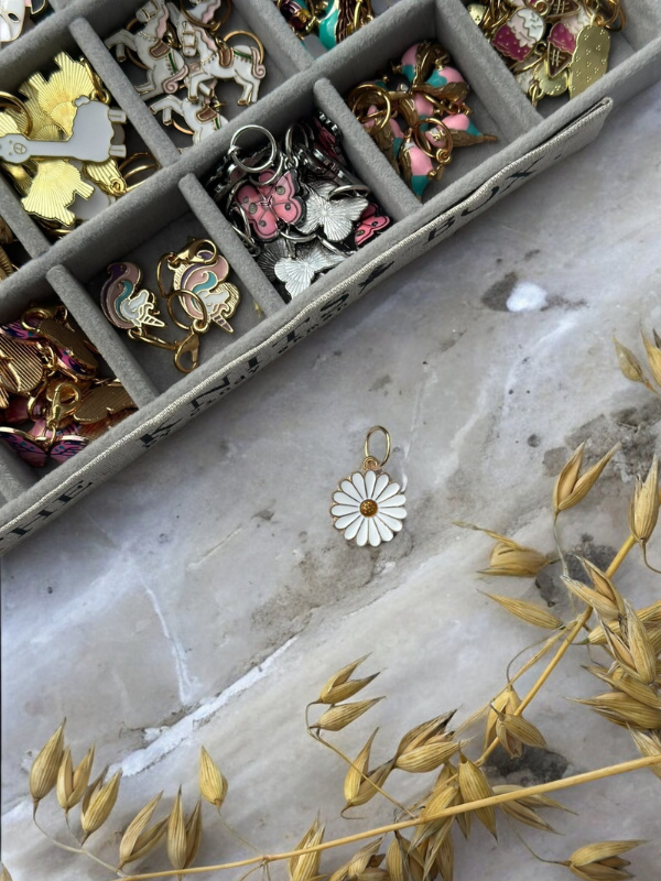A tray of KNITS by cindy ekman silmukkamerkit—colorful enamel stitch markers—rests on marble, with a white daisy charm and dried yellow grasses nearby, perfect for your neulonta projects.