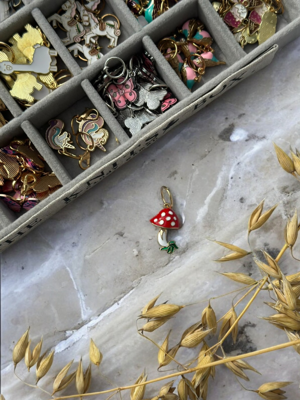 A KNITS by cindy ekman silmukkamerkit red and white mushroom charm with a green stem rests on marble near a gold jewelry organizer filled with kauniit silmukkamerkit—perfect neulonta accessories. Dry wheat stalks lie at the bottom right.