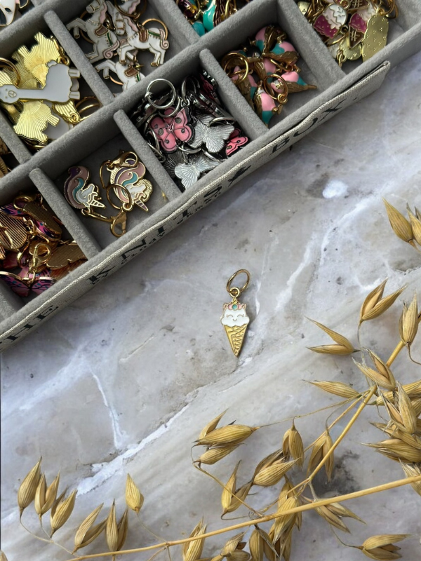 A KNITS by cindy ekman silmukkamerkit set sits in an organizer above a gold ice cream cone charm with white enamel on marble, while dried golden grasses decorate the lower right corner. Perfect for neulonta.