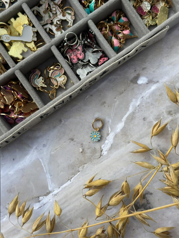 A KNITS by cindy ekman silmukkamerkit jewelry organizer tray with colorful silmukkamerkit rests on marble. In front, a single blue flower-shaped charm for neulonta is placed near dried golden grass.