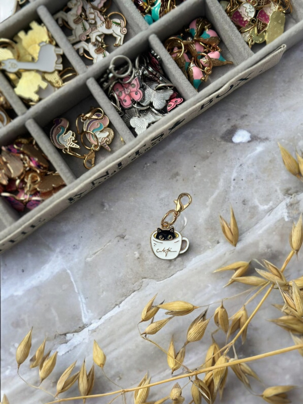 A tray of KNITS by cindy ekman silmukkamerkit sits on marble. Up front, a white cup-shaped charm with a black cat face and "café" is displayed for neulonta, surrounded by dried wheat stems.