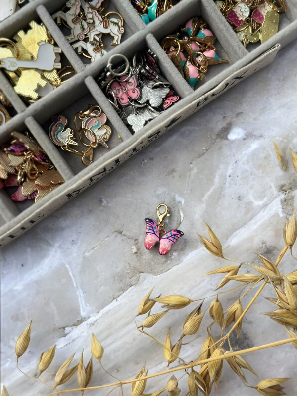 A tray displays organized, colorful KNITS by cindy ekman silmukkamerkit—mainly butterfly and floral shapes—on marble. A separate pink butterfly marker lies below near dried golden grass, perfect for your neulonta projects.