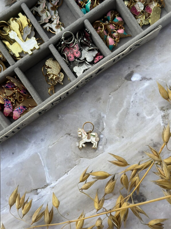 A display box features colorful animal enamel pins. Nearby, a gold and white KNITS by cindy ekman silmukkamerkit llama pin—ideal for neulonta—rests on the marble surface next to dried grass.