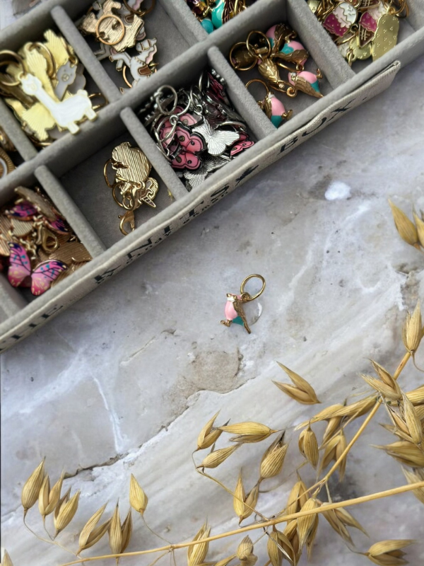 A gold and pink bird-shaped earring lies on a marble surface, with KNITS by cindy ekman silmukkamerkit in a colorful enamel and gold jewelry box in the upper left corner and dried oat stalks in the lower right.
