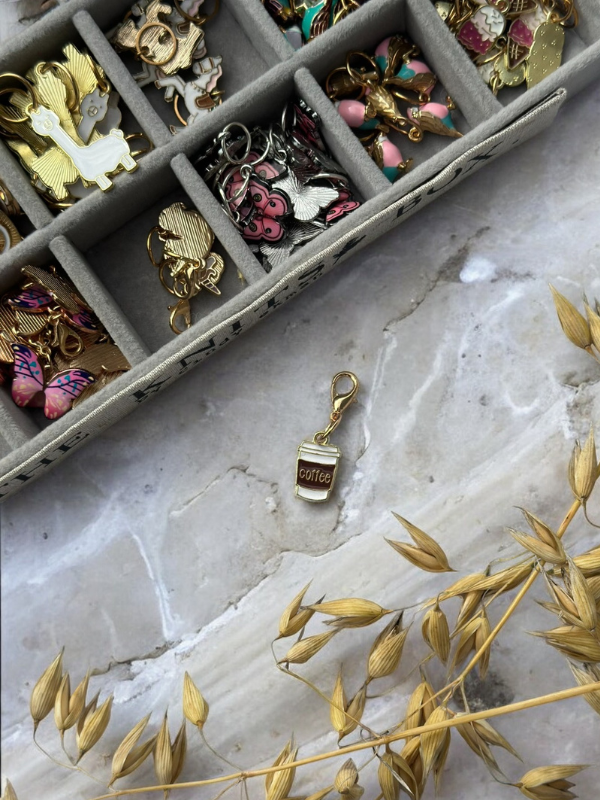 A jewelry box with colorful KNITS by cindy ekman silmukkamerkit for neulonta sits on a marble surface. In front is a gold coffee cup charm labeled “coffee,” with dried plants on the lower right side of the image.