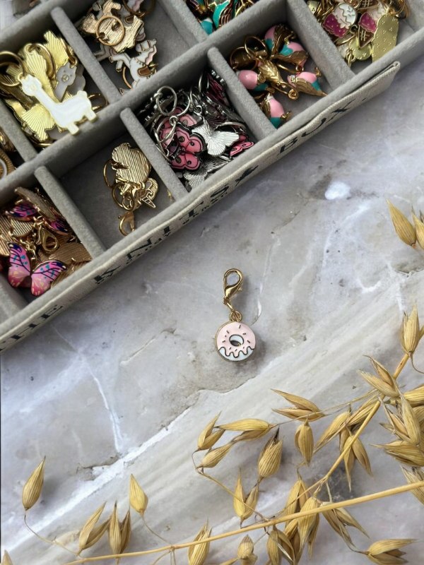 A tray of colorful KNITS by cindy ekman silmukkamerkit is displayed on a marble surface, with a pink frosted donut charm featuring a smiley face beside it, bringing playful charm to your neulonta essentials near some dried grass.
