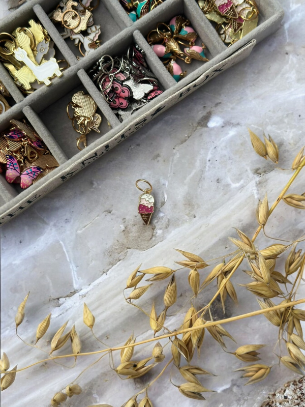 A jewelry box filled with KNITS by cindy ekman silmukkamerkit rests on a marble surface. A single pink and gold silmukkamerkki lies outside the box, next to dried golden grass.