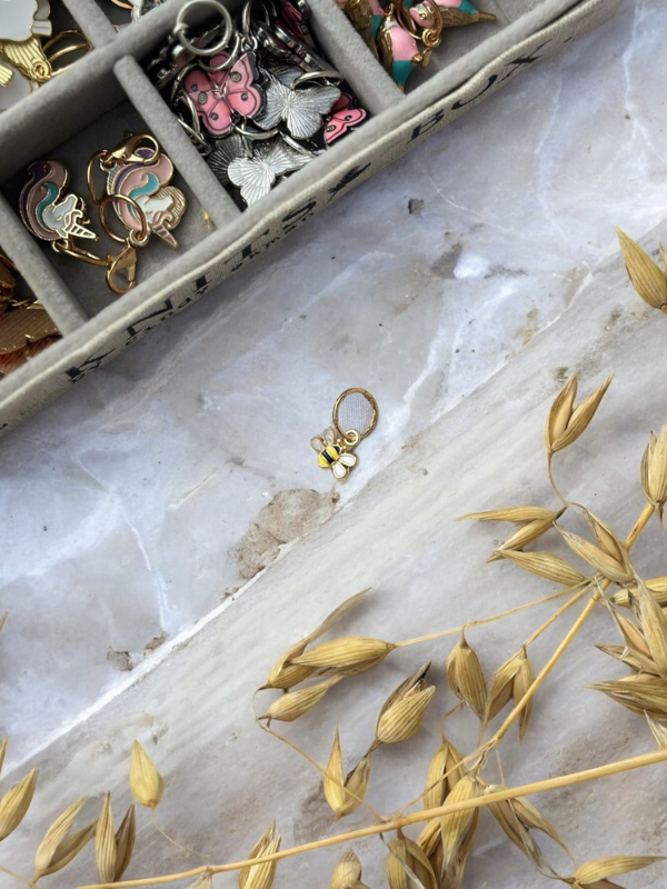 A close-up of a tray with assorted colorful KNITS by cindy ekman silmukkamerkit, a bee-shaped pin on marble, and dry wheat stalks—perfect inspiration for neulonta lovers.