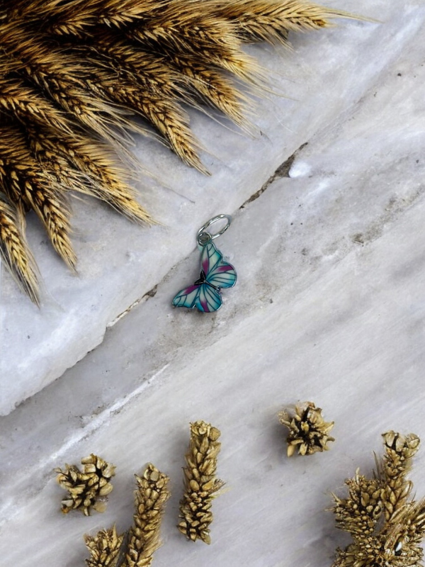 A KNITS by cindy ekman silmukkamerkit, a blue and purple butterfly-shaped pendant ideal for neulonta, rests on white marble, encircled by golden wheat stalks and small wheat clusters.