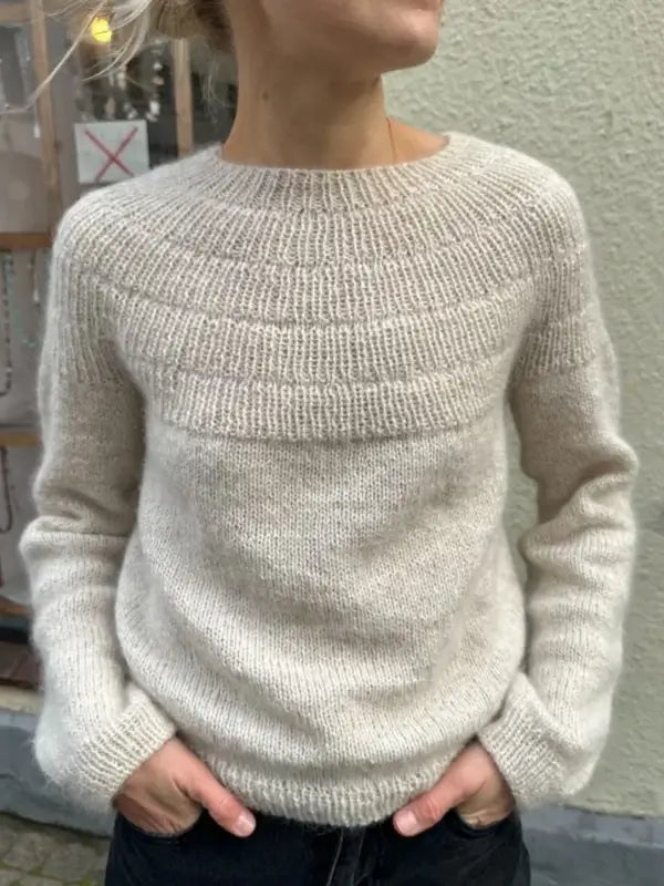 A person wearing the PetieKnit Anker’s Sweater - My Size neuleohje - Suomi in a light beige shade, featuring hand-knit textured details on the yoke, stands with hands in pockets against a light-colored wall. The person’s face is not visible.