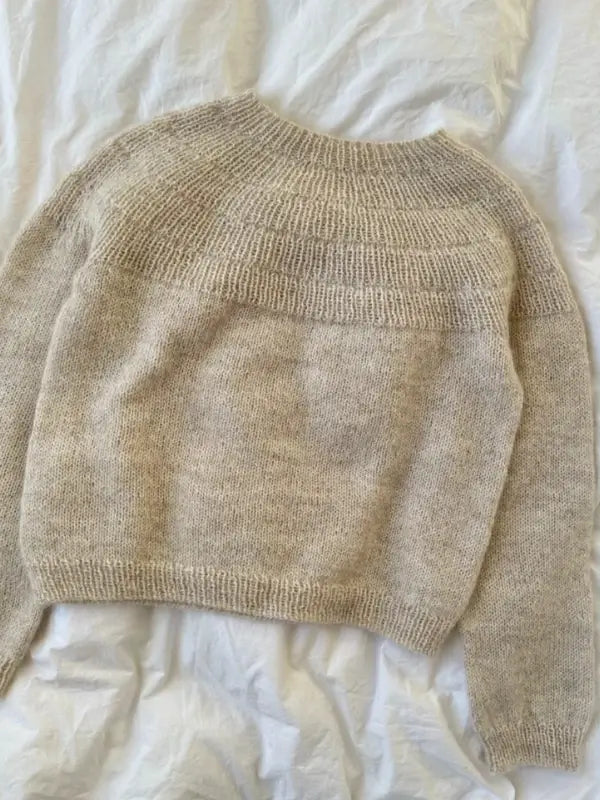 A beige, long-sleeved, knitted sweater is laid flat on a white, crumpled fabric background. The PetieKnit Anker’s Sweater – My Size neuleohje – Suomi features a round neckline and a textured yoke with ribbed cuffs, hem, and collar; the perfect inspiration if you’re searching for a neuleohje or suomenkielinen printti.