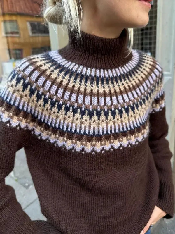 Wearing the PetiteKnit - Celeste Sweater neuleohje in brown, featuring a kirjoneule yoke patterned with white, beige, black, and light purple accents, a person stands outside near a building.