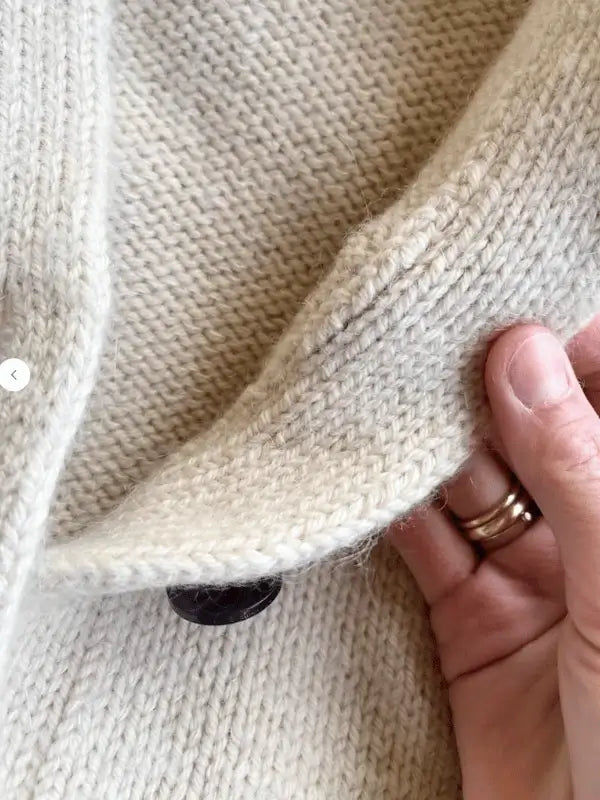 A close-up of a hand with rings holding the edge of an oversize cream-colored Champagne Cardigan, knitted using the PetiteKnit - Champagne Cardigan neuleohje, with a black button partially hidden beneath the fold.