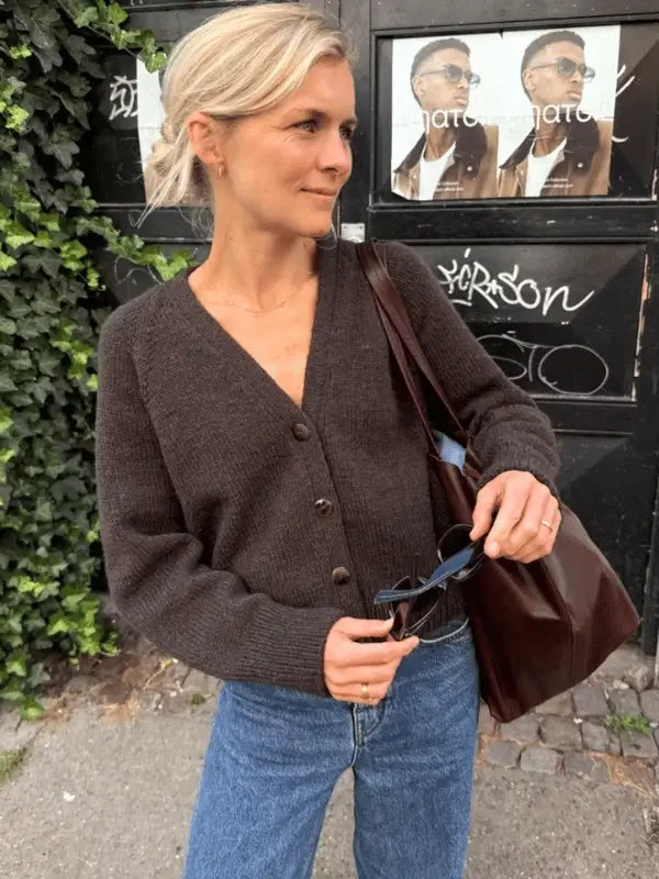 A woman with blonde hair in a loose bun wears the PetiteKnit - Champagne Cardigan neuleohje, blue jeans, and holds sunglasses and a brown tote bag while standing outdoors by a wall covered with posters and graffiti.