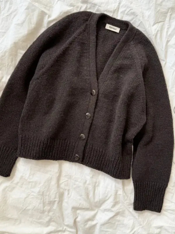 The PetiteKnit - Champagne Cardigan neuleohje, a dark brown oversized long-sleeve cardigan, is laid flat on a wrinkled white fabric background.