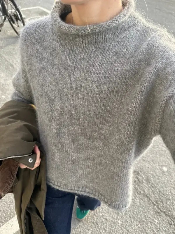 A person wearing a loose gray knit PetiteKnit - Cloud Sweater Neuleohje and blue jeans, holding a brown jacket. The outdoor photo is taken from above, showing only the upper body and part of the street.