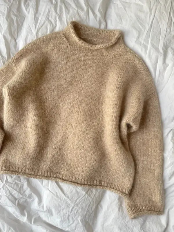 A beige, long-sleeve knitted sweater—PetiteKnit - Cloud Sweater Neuleohje—with a round neckline is laid flat on a white, crumpled fabric background, highlighting its elegant neuleohje design.