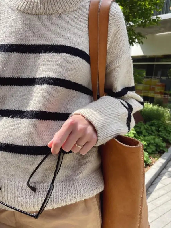 A person wearing the PetiteKnit Marseille Sweater neuleohje- Suomi holds eyeglasses and a tan shoulder bag while standing outdoors near greenery.