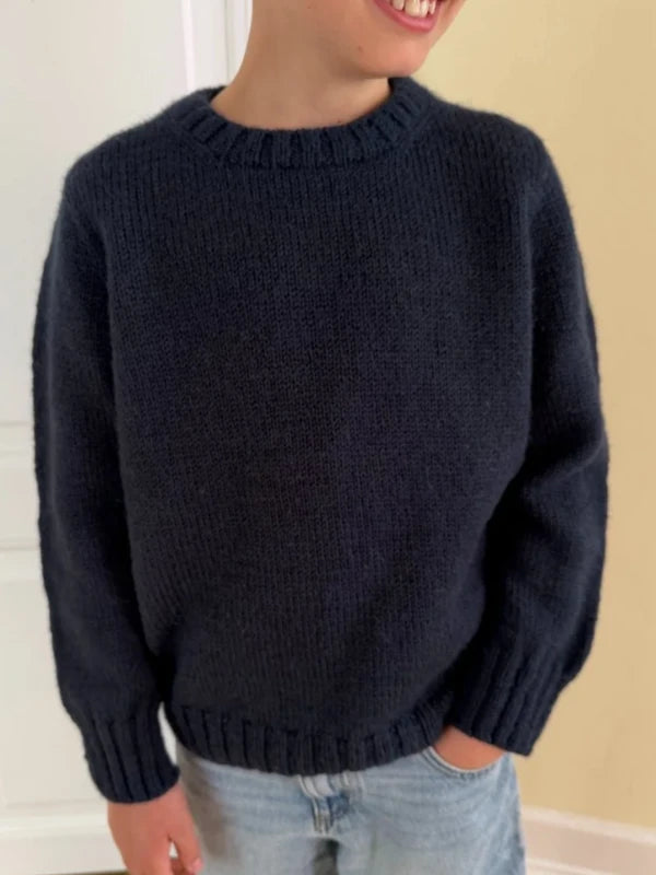A child wearing a dark blue PetiteKnit Niels Sweater Junior neuleohje, Suomi and light blue jeans stands against a light-colored wall, smiling and looking to the side.