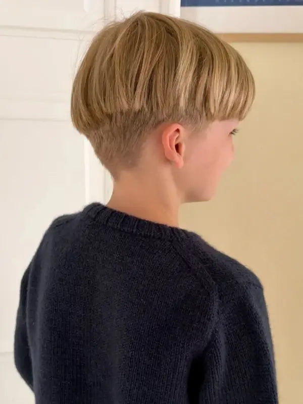 A young child with light blond hair in a bowl cut, wearing a dark blue PetiteKnit Niels Sweater Junior neuleohje (Suomi), is shown from behind and slightly to the side, standing indoors near a light-colored wall.