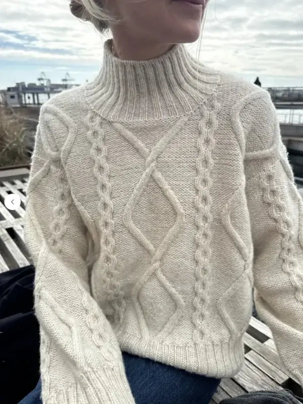 A person sits outdoors on a wooden bench, wrapped in a thick, cream-colored cable knit sweater with a mock neck—a cozy look crafted from the PetiteKnit Saga sweater neuleohje Suomi. With cloudy skies overhead and water in the background, the scene perfectly embodies the warmth and comfort inspired by this suomenkielinen neuleohje.