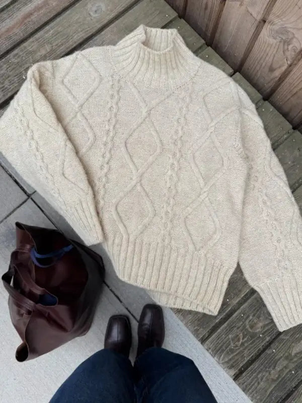 A cream-colored cable knit sweater, inspired by the PetiteKnit Saga sweater neuleohje Suomi, is laid out on a wooden bench next to a brown bag. In front stands a person in blue jeans and brown shoes, evoking classic Scandinavian style and showcasing suomenkielinen craftsmanship.