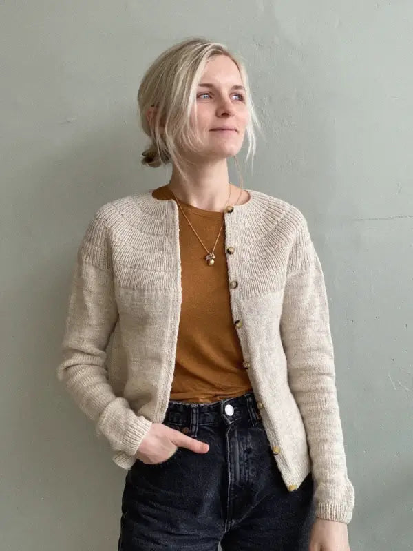A woman with blonde hair in a low bun wears the PetiteKnit - Anker's Cardigan MY SIZE neuleohje, a beige knit with a circular yoke, over a brown shirt and black jeans, standing against a light gray wall, smiling slightly.